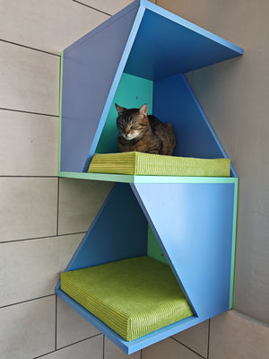 Double corner tower lounge shelf. The shelf is mounted onto a wall. One side of the wall is tiled while the other is regular. The tower is bright, colored in heaven blue with mint middle and back plates. It mounts onto both sides of the corner, making it extra stable. Shelf is modern with a twist. There is a tabby cat snoozing on a comfortable pillow. The pillow is thick and covered in lush velvet fabric with stripes, so called corduroy. It's bright green.