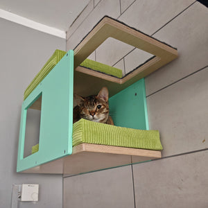 One eyed tabby cat is lying on a bright green cushion inside a wall mounted cat tunnel shelf. The cat shelf is mounted onto a tiled grey wall. The shelf is sturdy, made out of birch plywood. The front and back accent plates are mint colored, while the base top and bottom plates are in lacquered birch. The corner cat tunnel has one window and multiple entry points. There are 2 comfortable cushions, one on top, the other inside, both bright green corduroy
