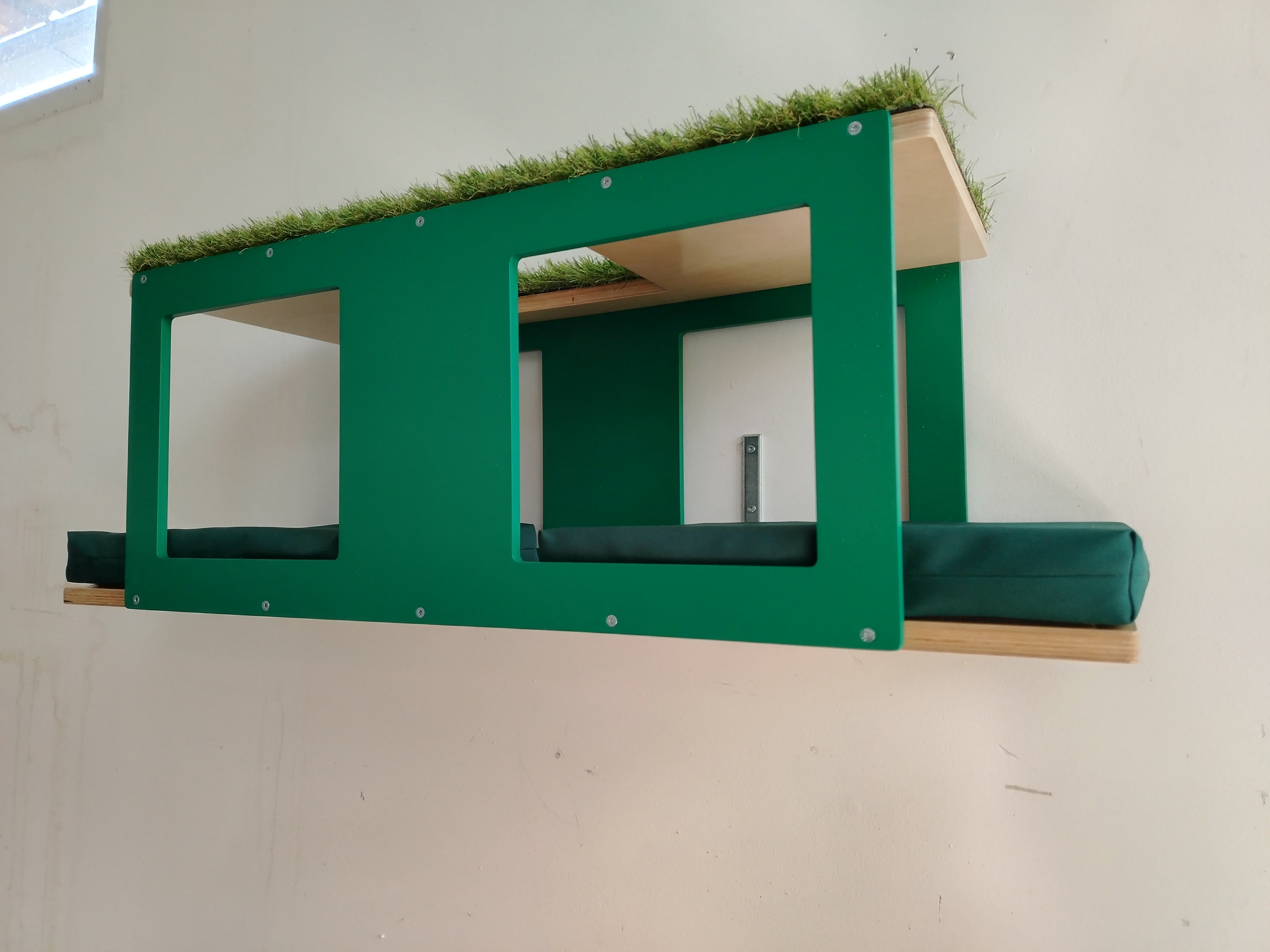 Outdoor enclosure mesh mounted big cat shelf. It's a multilevel platform with multiple entry points and windows. There are 2 comfortable cushions on the lower level. The top level is carpeted with artificial grass. The base color is natural, while the accent is emerald green. The cushions are in tenda green. 